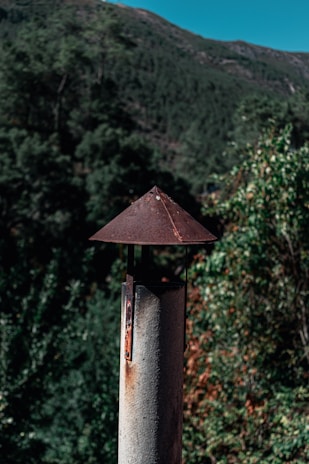 A rustic metal chimney pipe stands in the foreground, topped with a conical cap that shows signs of rust. The background features a lush forested landscape and a clear blue sky peeking over the top of a green mountain, suggesting a serene natural setting.