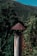 A rustic metal chimney pipe stands in the foreground, topped with a conical cap that shows signs of rust. The background features a lush forested landscape and a clear blue sky peeking over the top of a green mountain, suggesting a serene natural setting.