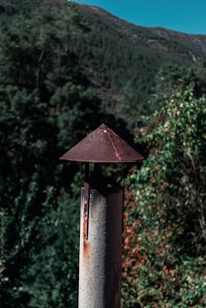 A rustic metal chimney pipe stands in the foreground, topped with a conical cap that shows signs of rust. The background features a lush forested landscape and a clear blue sky peeking over the top of a green mountain, suggesting a serene natural setting.