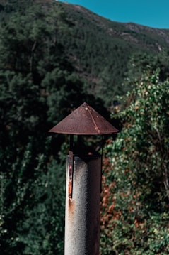 A rustic metal chimney pipe stands in the foreground, topped with a conical cap that shows signs of rust. The background features a lush forested landscape and a clear blue sky peeking over the top of a green mountain, suggesting a serene natural setting.