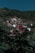 A cluster of rustic houses with stone facades is nestled on a lush, green hillside. The roofs of the houses vary in color, with some being red and others a darker shade. Dense forest surrounds the village, creating a serene and secluded atmosphere.