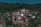 Stone houses are nestled on a lush green hillside, surrounded by dense forest. The structures appear rustic, with dark, earthy tones that blend with the natural environment. Small paths weave through the village, suggesting a remote, peaceful setting.
