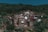 Stone houses are nestled on a lush green hillside, surrounded by dense forest. The structures appear rustic, with dark, earthy tones that blend with the natural environment. Small paths weave through the village, suggesting a remote, peaceful setting.