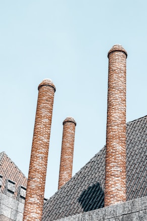 Several tall, cylindrical brick chimneys extend from a tiled rooftop. The bricks are arranged in a uniform, spiral pattern. The roof features a rustic, brownish tile design, contrasting against a clear, blue sky.