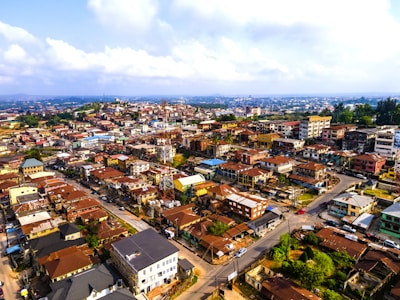 An aerial view of a densely populated urban area with a variety of buildings, including residential and commercial properties. The rooftops are predominantly reddish-brown, and the layout of the streets suggests a network of roads and intersections. In the background, a sprawling cityscape stretches into the distance, bordered by a partly cloudy sky.