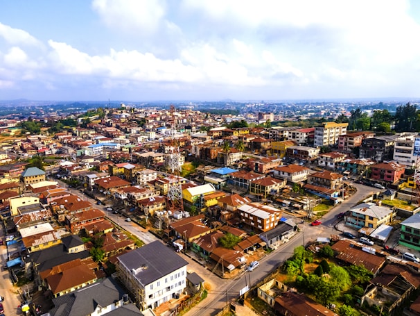 An aerial view of a densely populated urban area with a variety of buildings, including residential and commercial properties. The rooftops are predominantly reddish-brown, and the layout of the streets suggests a network of roads and intersections. In the background, a sprawling cityscape stretches into the distance, bordered by a partly cloudy sky.