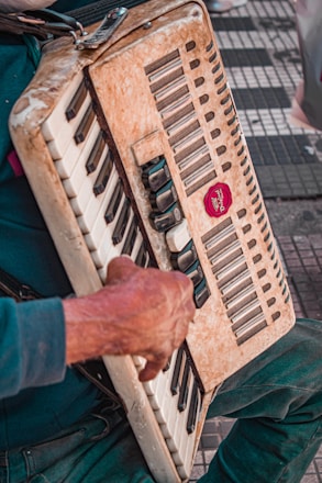 An aged accordion is held by a pair of hands, positioned on the instrument's keys and buttons. The surface of the accordion shows signs of wear, adding a vintage feel. The scene is set on a tiled or checkered ground, possibly outdoors.