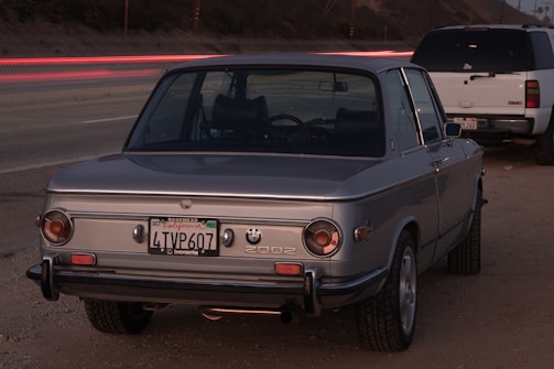 A vintage silver BMW 2002 car is parked on a dirt roadside. The vehicle's rear view is prominently shown, with its license plate visible. There is a white SUV parked nearby. The scene features blurred, long exposure lights from passing vehicles on the highway in the background, creating streaks of red and white.