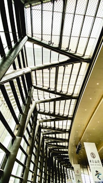 The image depicts a modern architectural interior, featuring tall steel pillars and a high glass ceiling with a grid-like pattern. There are large banners hanging from the ceiling, one of which displays the logo and name of Bank of China.