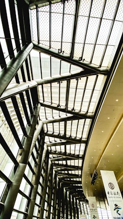 The image depicts a modern architectural interior, featuring tall steel pillars and a high glass ceiling with a grid-like pattern. There are large banners hanging from the ceiling, one of which displays the logo and name of Bank of China.