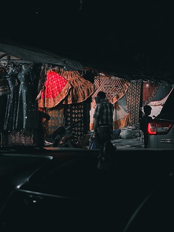 A dimly lit street market with colorful textiles prominently displayed. Vibrant fabrics in shades of red, gold, and orange hang from the stall, showcasing intricate patterns. A silhouette of a person in casual clothing stands near the textiles, while a silver vehicle is partially visible in the foreground, and a motorbike is parked nearby.