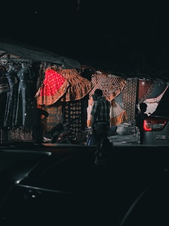 A dimly lit street market with colorful textiles prominently displayed. Vibrant fabrics in shades of red, gold, and orange hang from the stall, showcasing intricate patterns. A silhouette of a person in casual clothing stands near the textiles, while a silver vehicle is partially visible in the foreground, and a motorbike is parked nearby.