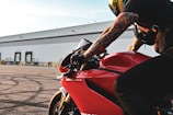 A motorcyclist with tattooed arms is leaning forward on a red Ducati 1199 Panigale motorcycle. The background features an industrial building with multiple loading docks. The image captures the details of the motorcycle and the rider's gear, including a helmet and gloves.