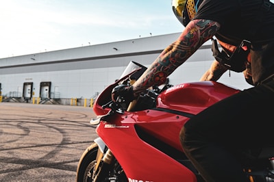 A motorcyclist with tattooed arms is leaning forward on a red Ducati 1199 Panigale motorcycle. The background features an industrial building with multiple loading docks. The image captures the details of the motorcycle and the rider's gear, including a helmet and gloves.