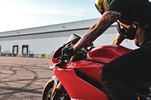 A motorcyclist with tattooed arms is leaning forward on a red Ducati 1199 Panigale motorcycle. The background features an industrial building with multiple loading docks. The image captures the details of the motorcycle and the rider's gear, including a helmet and gloves.