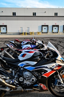Several high-performance motorcycles are parked in a row in front of an industrial building with loading docks. The bikes are sleek, featuring a prominent racing design with a combination of red, white, and blue colors. The building in the background has a flat roof and multiple garage-style doors.