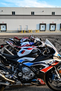 Several high-performance motorcycles are parked in a row in front of an industrial building with loading docks. The bikes are sleek, featuring a prominent racing design with a combination of red, white, and blue colors. The building in the background has a flat roof and multiple garage-style doors.