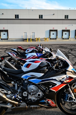 Several high-performance motorcycles are parked in a row in front of an industrial building with loading docks. The bikes are sleek, featuring a prominent racing design with a combination of red, white, and blue colors. The building in the background has a flat roof and multiple garage-style doors.