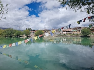 Tibetan prayer flags stretch over a calm, reflective body of water with lush greenery lining the shores. Traditional buildings are visible in the background beneath a sky filled with fluffy clouds.