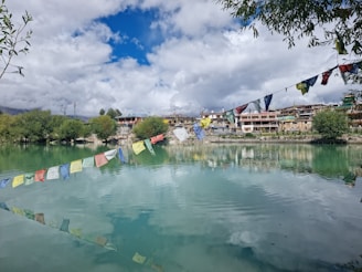 Tibetan prayer flags stretch over a calm, reflective body of water with lush greenery lining the shores. Traditional buildings are visible in the background beneath a sky filled with fluffy clouds.