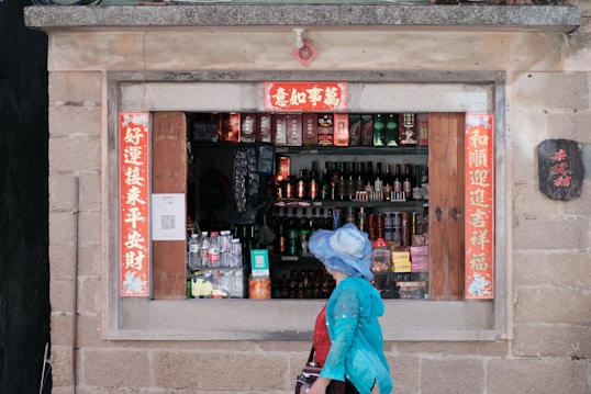 A small storefront with an open window displaying various bottles, cans, and boxes inside. The shop has traditional Chinese decorations with red banners containing Chinese characters. A person wearing a blue hat and blue jacket is walking past the shop window.