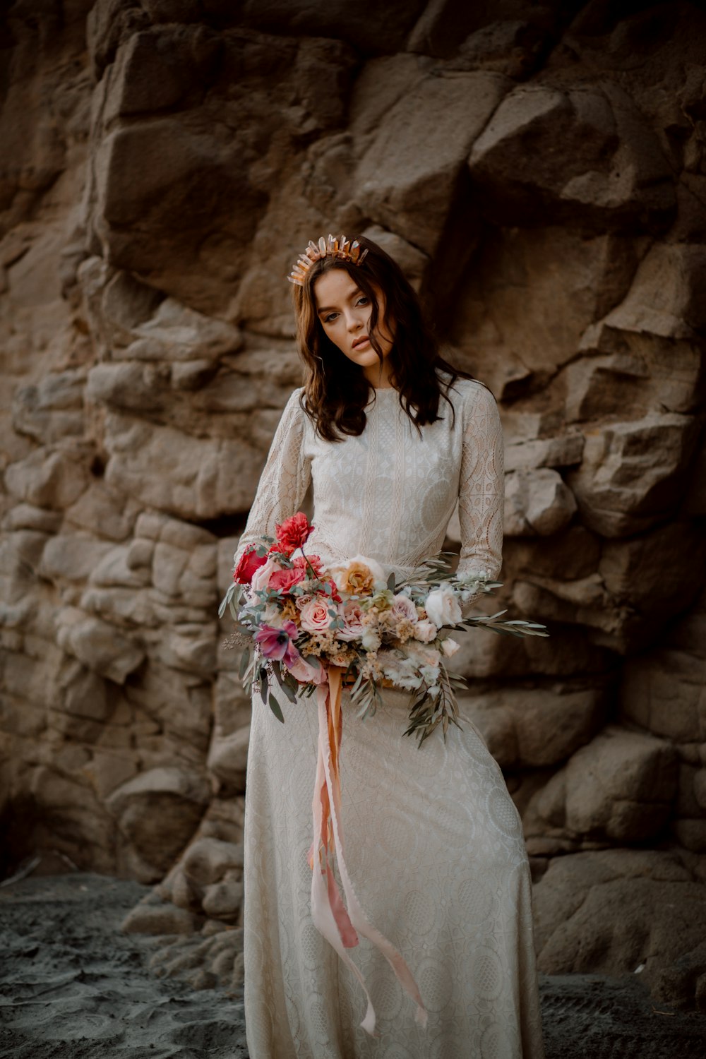 Una mujer con un vestido blanco con una corona y flores frente a una pared de roca