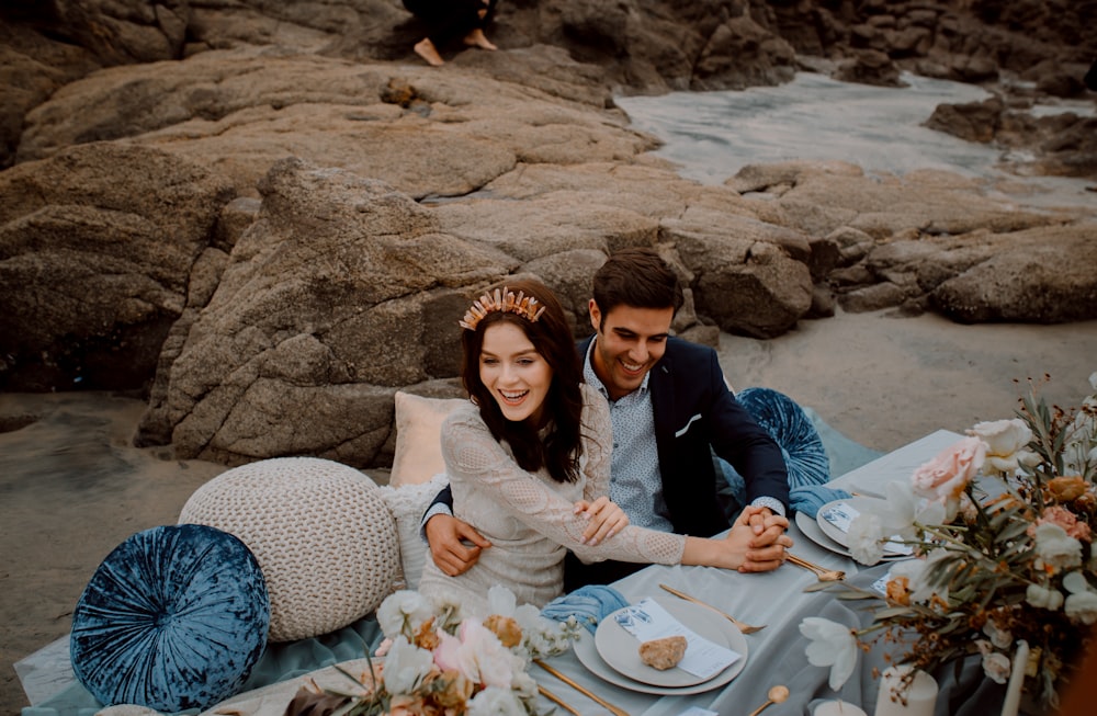 a man and woman sitting on a beach with food and flowers