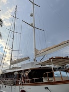A large sailing yacht with tall white masts and multiple sails, set against a backdrop of a cloudy sky with hints of sunlight. The yacht features a wooden deck and polished railings, with various ropes and rigging visible. Part of a palm tree is visible on the left side of the image.