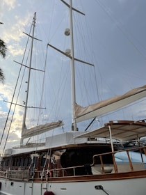 A large sailing yacht with tall white masts and multiple sails, set against a backdrop of a cloudy sky with hints of sunlight. The yacht features a wooden deck and polished railings, with various ropes and rigging visible. Part of a palm tree is visible on the left side of the image.