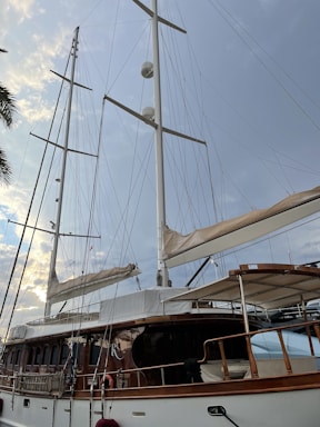 A large sailing yacht with tall white masts and multiple sails, set against a backdrop of a cloudy sky with hints of sunlight. The yacht features a wooden deck and polished railings, with various ropes and rigging visible. Part of a palm tree is visible on the left side of the image.