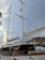 A large sailing yacht with tall white masts and multiple sails, set against a backdrop of a cloudy sky with hints of sunlight. The yacht features a wooden deck and polished railings, with various ropes and rigging visible. Part of a palm tree is visible on the left side of the image.