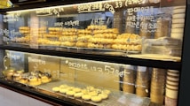 A display case filled with various baked goods including pastries, muffins, and pies. The display case features handwritten labels on the glass describing each item. There are also several stacks of paper cups and cones arranged on the side.