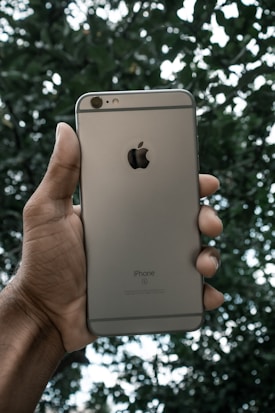 A hand is holding a smartphone with a metallic finish and an apple logo on the back, set against a background of green foliage.