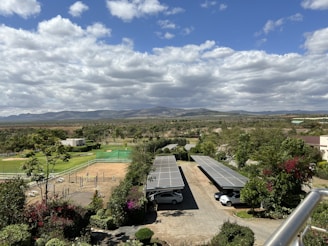 A scenic landscape featuring a mix of natural and man-made elements. In the foreground, there are two carports with several solar panels installed on their roofs and vehicles parked underneath. Surrounding the structures are lush green plants and colorful flowers. In the middle ground, more greenery and trees can be seen, along with a manicured lawn and some sports facilities, such as tennis courts. The background showcases sprawling open land and distant hills under a sky filled with clouds.
