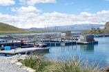 A series of rectangular fish farming platforms floating on a clear blue body of water, surrounded by grass and rocky shores. In the background, rolling hills and snow-capped mountains are visible under a partly cloudy sky, with power lines and parked vehicles nearby.