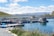 A series of rectangular fish farming platforms floating on a clear blue body of water, surrounded by grass and rocky shores. In the background, rolling hills and snow-capped mountains are visible under a partly cloudy sky, with power lines and parked vehicles nearby.