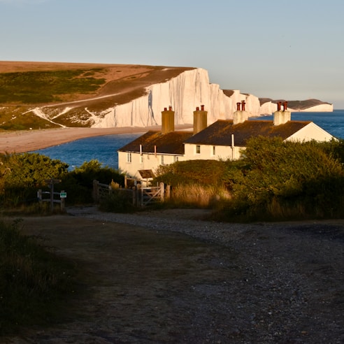 A coastal landscape with prominent white chalk cliffs along the shoreline, partially illuminated by sunlight. In the foreground, there are two quaint cottages with pitched roofs and visible chimneys, surrounded by lush green vegetation. The scene has a serene atmosphere, with a distant view of the blue sea meeting the horizon.