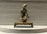 A small, intricate bronze sculpture of a kneeling figure mounted on a raised platform. The figure is detailed with traditional attire and a headpiece, appearing to represent a religious or cultural icon. The background consists of a textured gray surface that creates contrast with the metallic sheen of the sculpture.