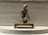 A small, intricate bronze sculpture of a kneeling figure mounted on a raised platform. The figure is detailed with traditional attire and a headpiece, appearing to represent a religious or cultural icon. The background consists of a textured gray surface that creates contrast with the metallic sheen of the sculpture.