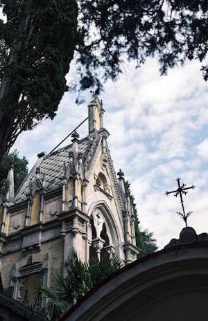 A gothic-style architectural structure with intricate details on its facade, surrounded by tall trees and a cloudy sky. The building features pointed arches, ornate windows, and stone embellishments.