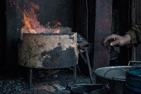 A metal container with a rugged, burnt surface holding a blazing fire emits vibrant flames. A person's hand reaches towards the fire, while an industrial setting with metal components surrounds the area.