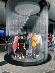A group of people is standing inside a circular indoor waterfall feature. Water cascades down from the ceiling around them, creating a unique and immersive experience. The individuals seem engaged, with some smiling and others taking photos. The setting appears to be a public space, possibly an aquarium or a science center.