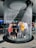 A group of people is standing inside a circular indoor waterfall feature. Water cascades down from the ceiling around them, creating a unique and immersive experience. The individuals seem engaged, with some smiling and others taking photos. The setting appears to be a public space, possibly an aquarium or a science center.