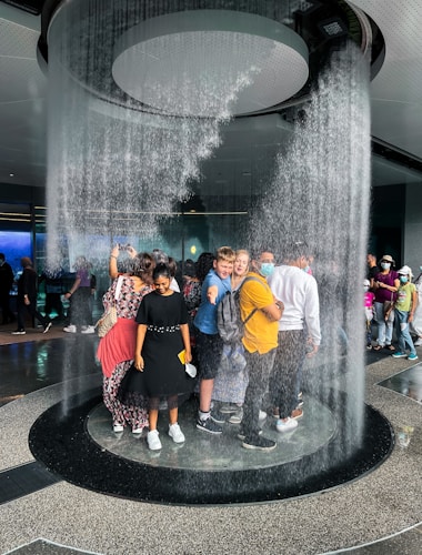 A group of people is standing inside a circular indoor waterfall feature. Water cascades down from the ceiling around them, creating a unique and immersive experience. The individuals seem engaged, with some smiling and others taking photos. The setting appears to be a public space, possibly an aquarium or a science center.