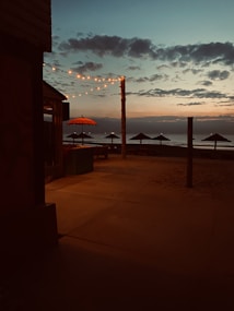 A tranquil beachside scene at dusk with string lights hanging from a building, casting a warm glow. The sky is a gradient from blue to orange, with scattered clouds. An umbrella with a bright, reddish canopy and several beach canopies are silhouetted against the ocean.