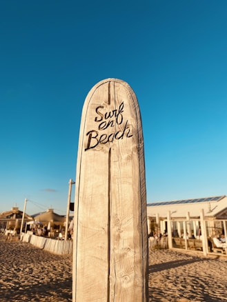 A wooden surfboard-shaped sign with the words 'Surf en Beach' carved into it stands upright on a sandy beach. In the background, there are buildings with large windows and people visible near the structures. The sky is clear and blue, indicating a sunny day.