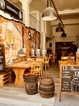 The interior of a rustic bar or brewery featuring wooden tables and chairs, two large metal pendant lights, barrels used as decoration, and a large wall mural depicting a bearded glass of beer. A chalkboard advertisement promotes 12 beers on tap, along with tastings, live music, events, and take-away beer. The overall ambiance is warm with brick walls and ample natural lighting.