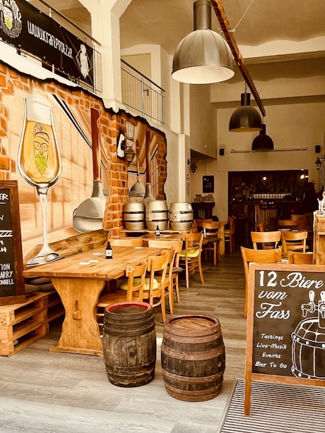 The interior of a rustic bar or brewery featuring wooden tables and chairs, two large metal pendant lights, barrels used as decoration, and a large wall mural depicting a bearded glass of beer. A chalkboard advertisement promotes 12 beers on tap, along with tastings, live music, events, and take-away beer. The overall ambiance is warm with brick walls and ample natural lighting.