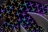 A pattern of colorful illuminated lines forms star-like shapes, set against a dark background. Curved metallic structures with a reflective surface intersect the pattern, creating a futuristic and vibrant appearance.