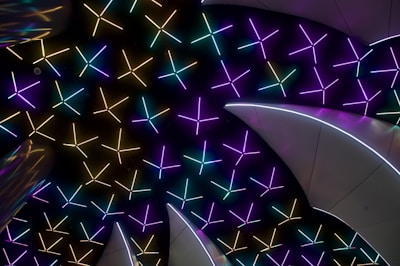 A pattern of colorful illuminated lines forms star-like shapes, set against a dark background. Curved metallic structures with a reflective surface intersect the pattern, creating a futuristic and vibrant appearance.